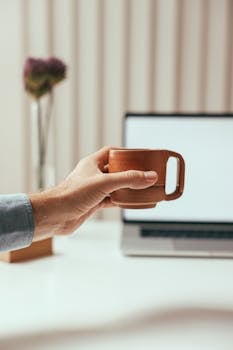 Close-up of a hand holding a coffee mug in a modern office setup with a laptop.