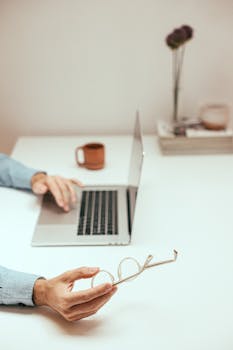 Clean workspace with a laptop, coffee cup, and hands holding glasses, perfect for productivity.