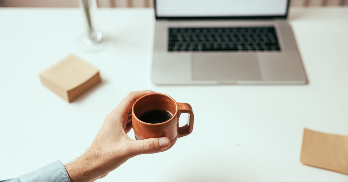A person holds a brown coffee cup near a laptop on a clean white desk, perfect for remote work themes. - Evidence evidence guide for caffeine focus randomized trial