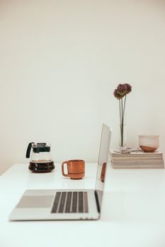 A serene minimalist workspace featuring a laptop, coffee pot, and flower vase on a white desk.