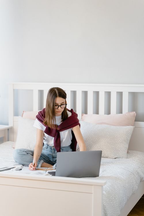 Free Woman sitting on bed with laptop, participating in remote learning session indoors. Stock Photo