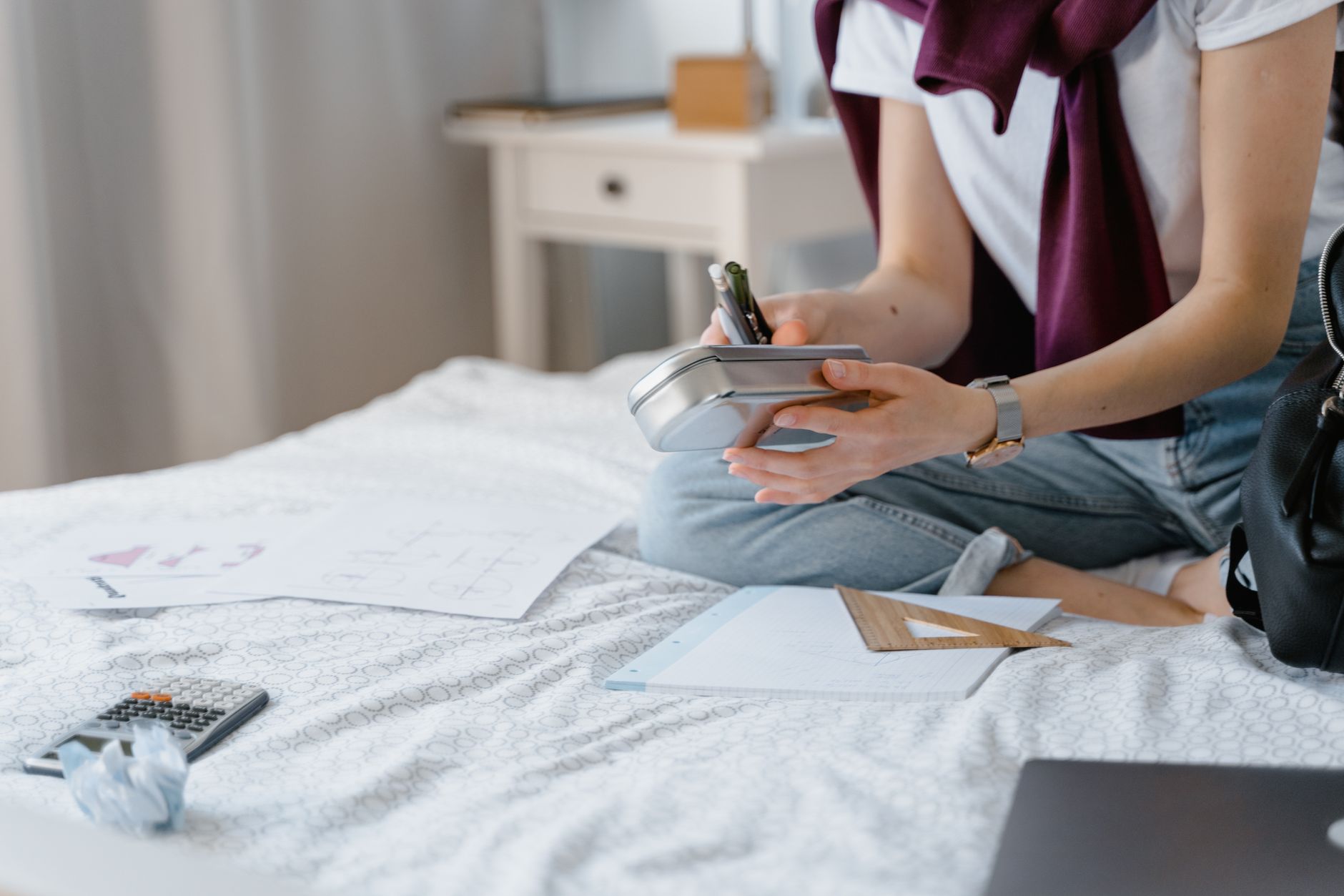 A student sitting on a bed while organizing study materials in a cozy bedroom environment.
