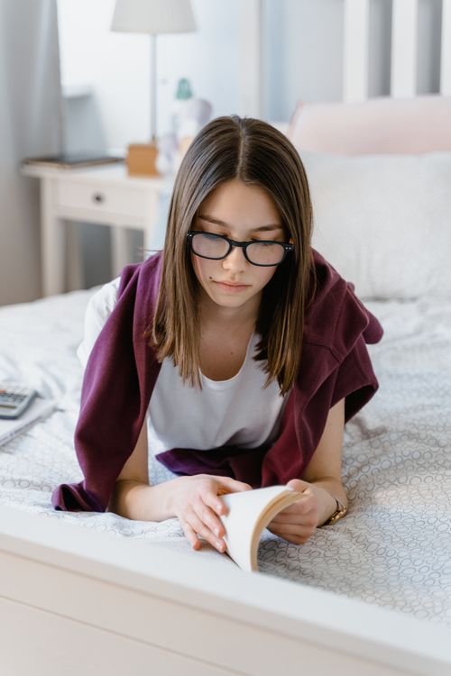 Free Teenage girl with eyeglasses reading a book in a cozy bedroom setting, focused and relaxed. Stock Photo