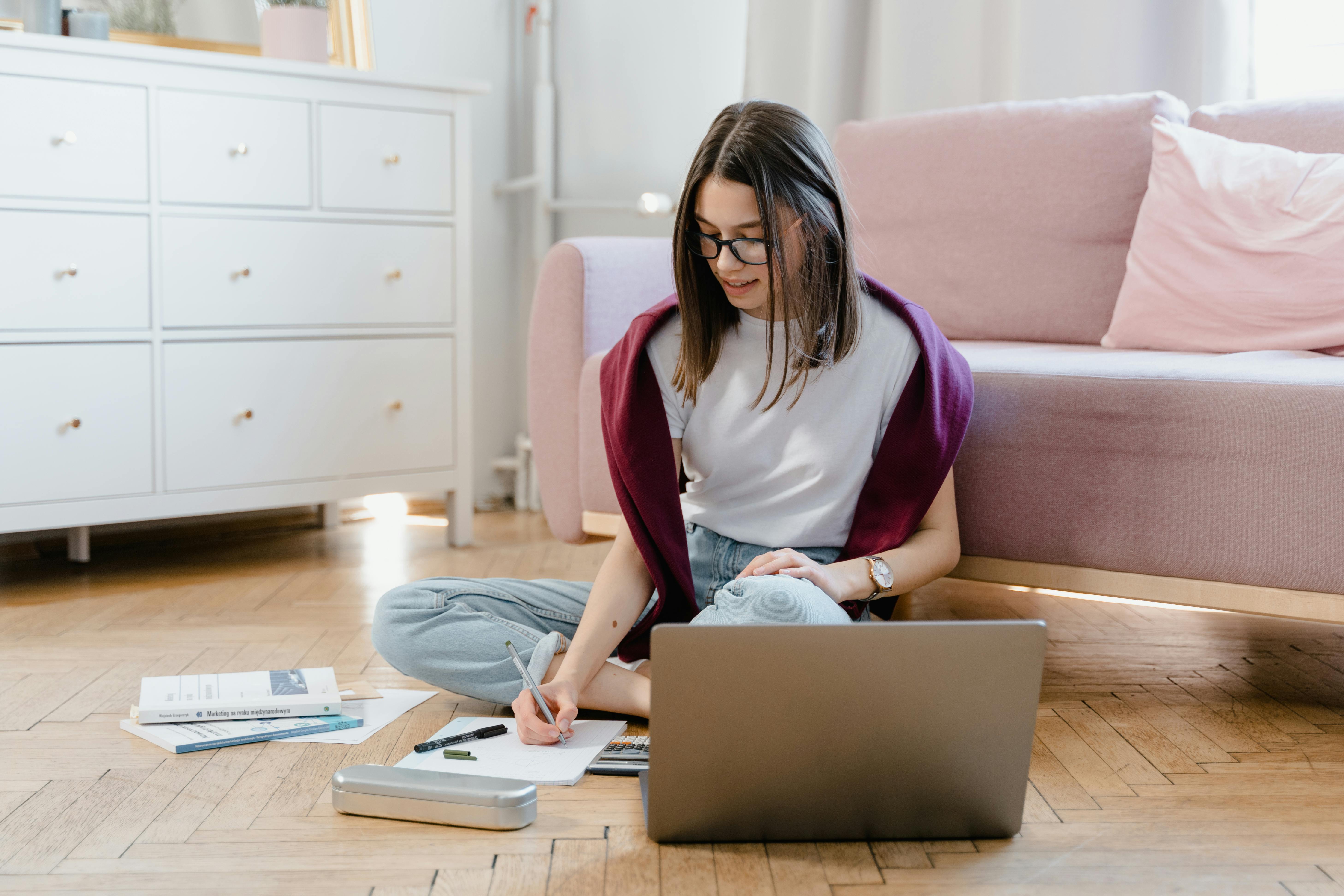 A young woman studying indoors using a laptop and writing notes.