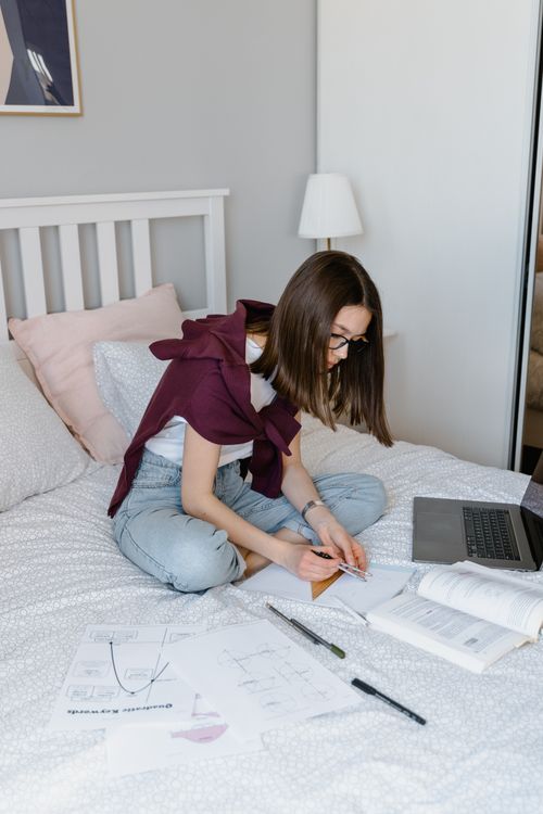 Free A young woman studying at home using a laptop, papers, and books on her bed. Stock Photo