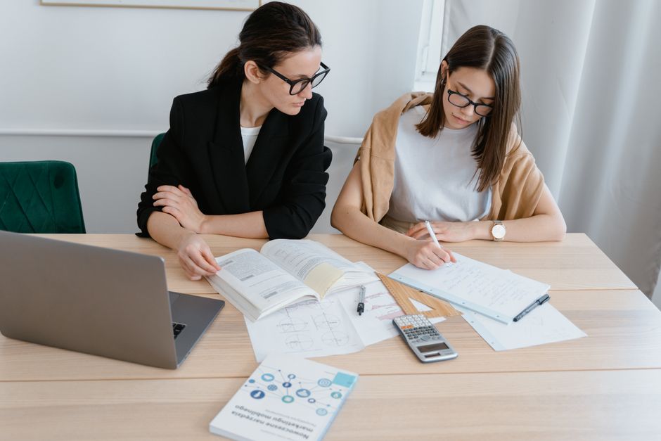 Two women at a desk studying with books, notes, and a laptop, emphasizing collaboration.