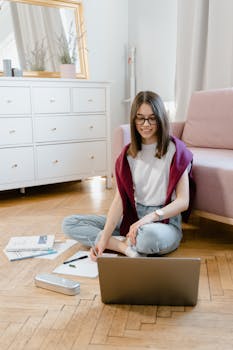 Teen girl engaged in online learning at home, writing notes with laptop on the floor.
