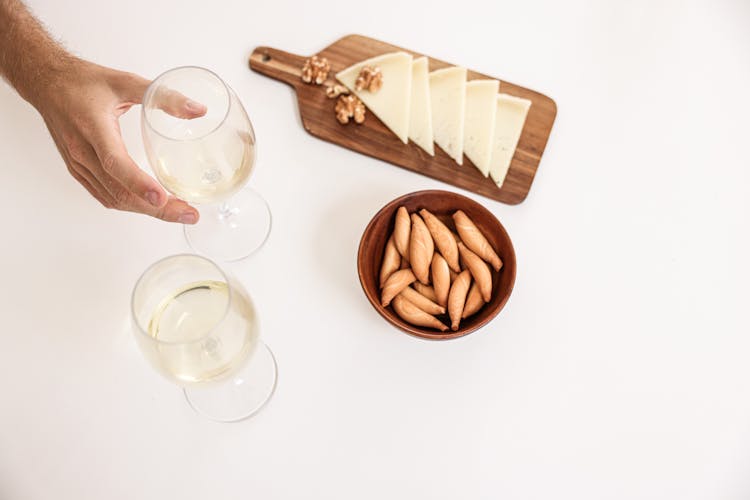 A Person Holding A Glass Of Wine Near The Cheese On A Wooden Board