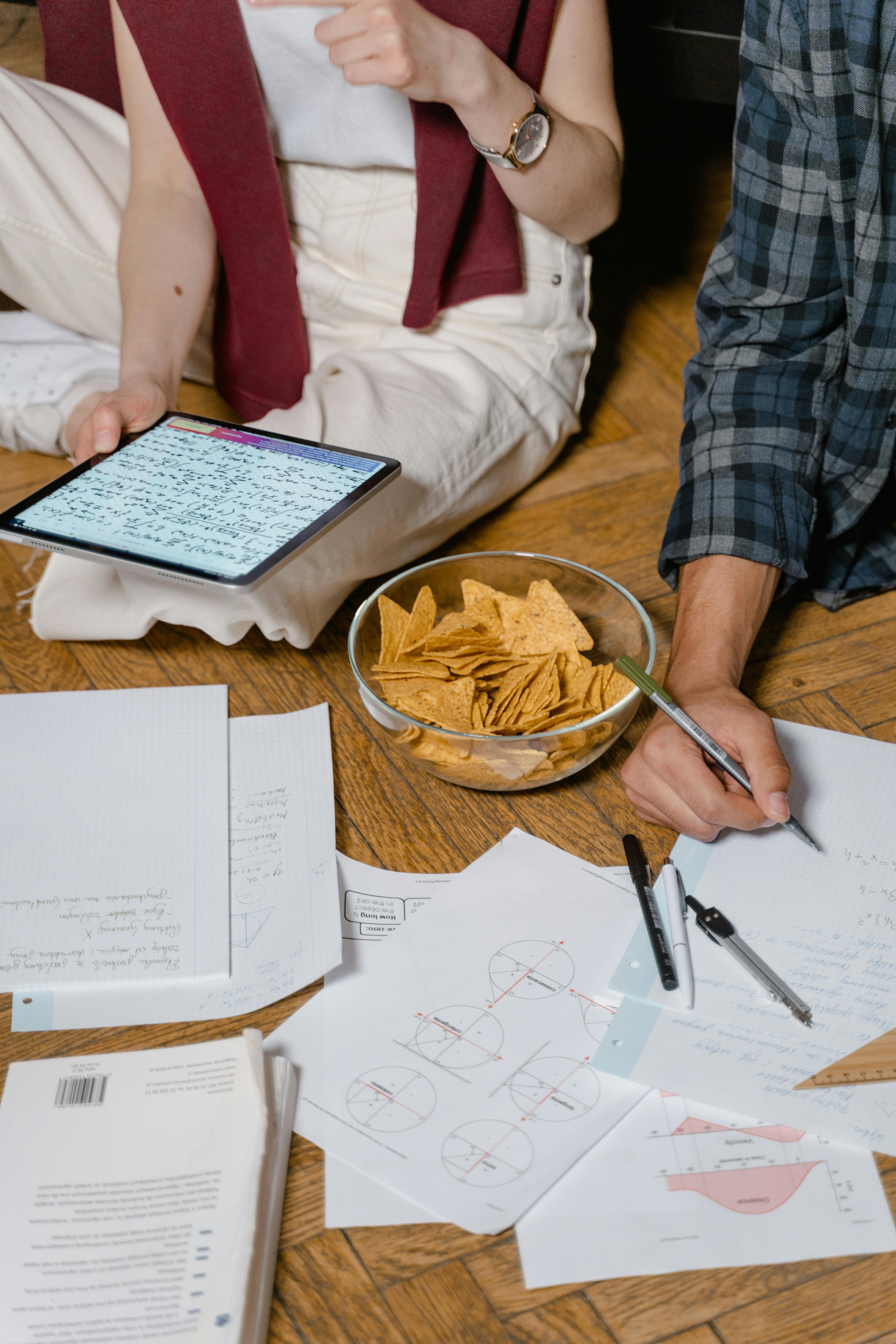 a person in plaid long sleeves writing on paper near the person in white pants holding a tablet