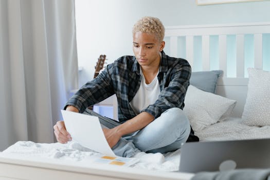 Young man studying on bed with laptop and notes, focused on remote learning indoors.