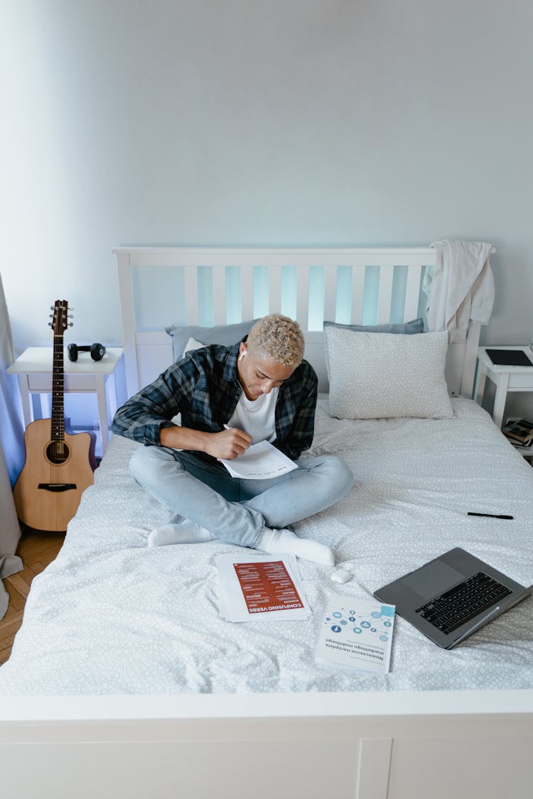 A Man In Plaid Long Sleeves Sitting On The Bed While Writing On Paper