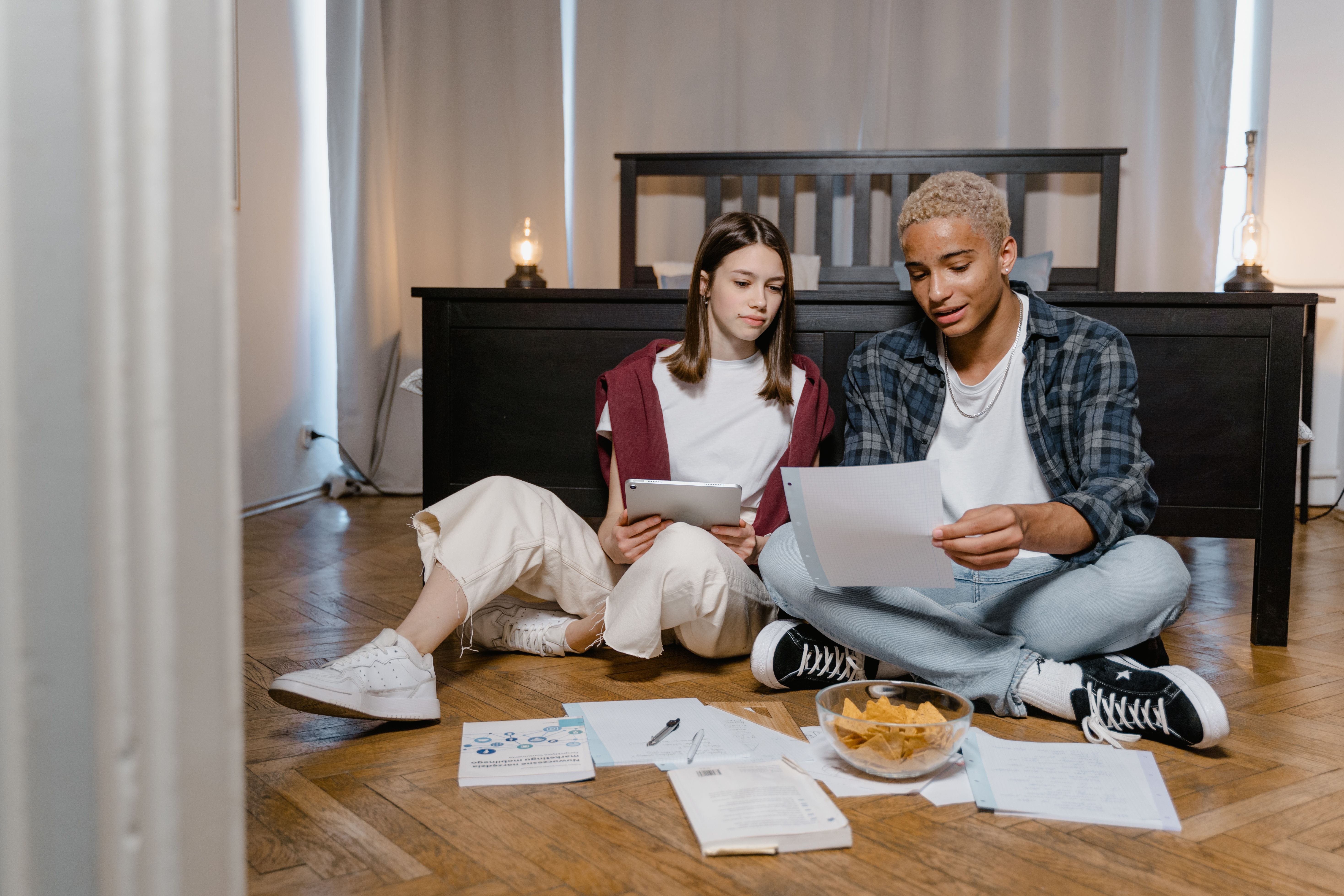 Two young adults studying with a tablet and papers on a bedroom floor.