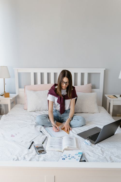 Free A young woman studying on a bed with a laptop, glasses, and books in a bright bedroom setting. Stock Photo