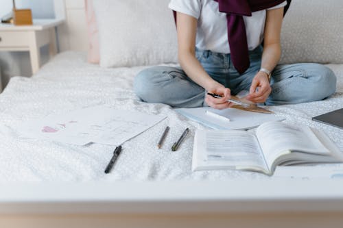 A Person in White Shirt and Denim Jeans Sitting on the Bed while Holding a Pen