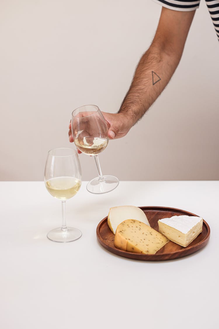 A Person Holding A Wine Glass Next To A Selection Of Cheese