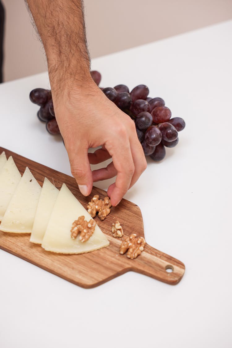 A Person Holding A Walnut On A Wooden Board With Sliced Cheese