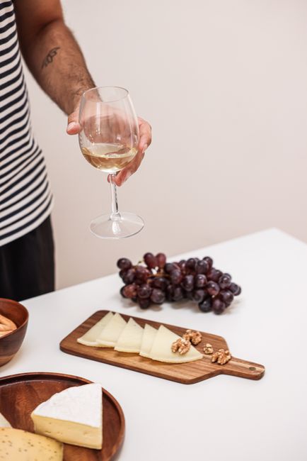 Person holding a wine glass over a wooden board with assorted cheeses on a white table.