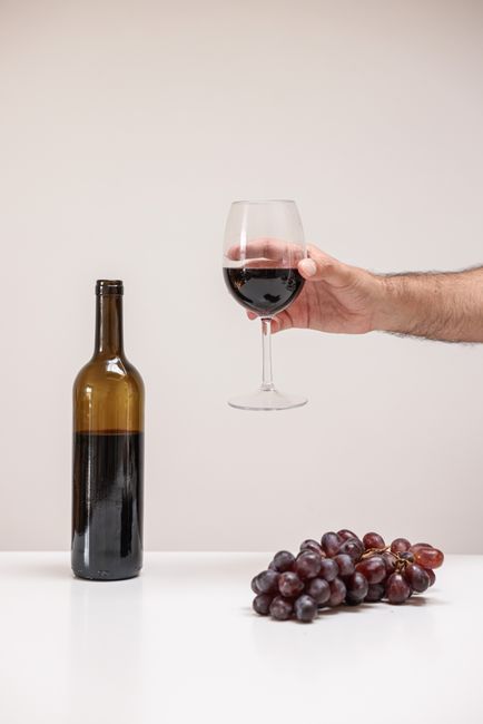 A hand holding a glass of red wine next to a bottle and fresh grapes on a white table.
