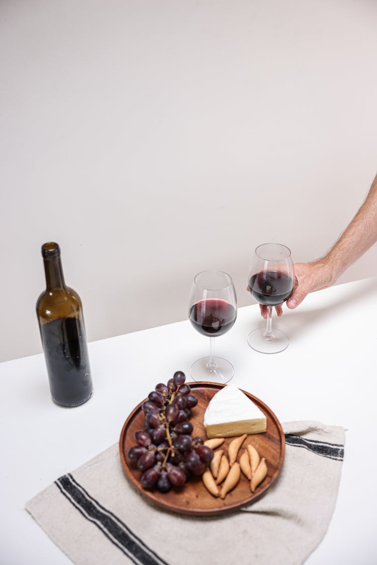 A Person Holding A Glass Of Wine Near The Wooden Plate With Grapes And Cheese