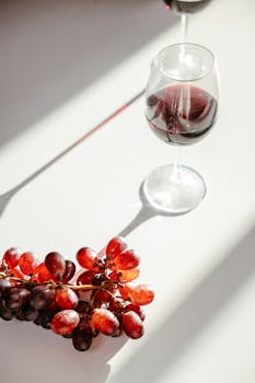 Red wine glass and grapes casting shadows on a sunlit table, creating a warm and inviting atmosphere.