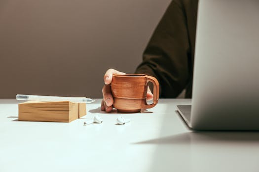 Person holding coffee mug at a minimalistic office desk with laptop, notepad, pen, and earbuds.