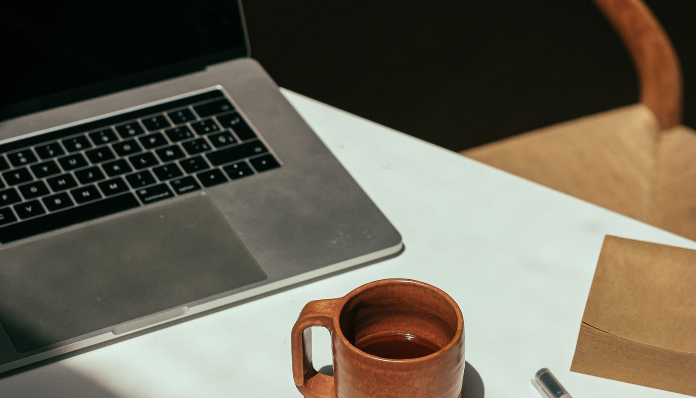 An open laptop with a darkened screen beside a terracotta mug and a pen on a sunlit desk