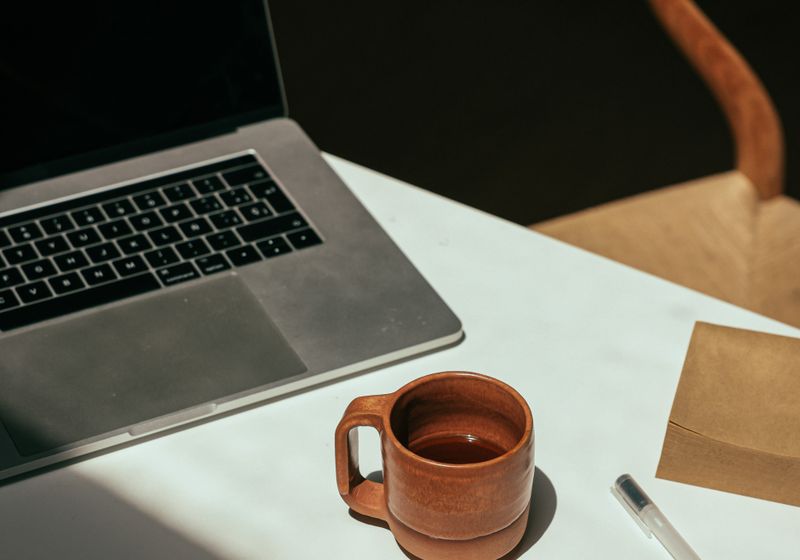 An open laptop with a darkened screen beside a terracotta mug and a pen on a sunlit desk