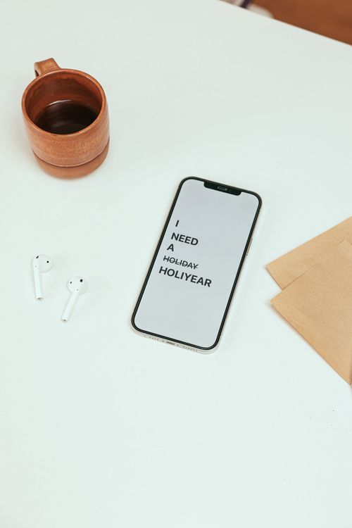 Free Smartphone displaying 'I need a holiday' text on a clean desk with coffee and earphones. Stock Photo
