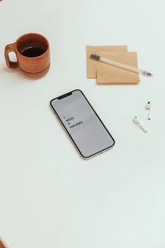 A minimalist desk setup with a smartphone showing 'I NEED A HOLIDAY,' coffee cup, AirPods, and stationery.
