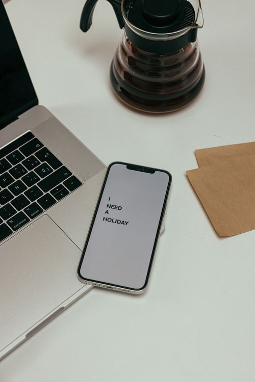 Free A smartphone on a desk displaying the message 'I need a holiday' next to a laptop and coffee pot. Stock Photo