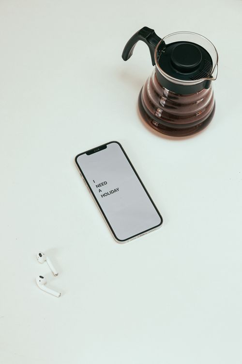 Free Flat lay of a smartphone displaying 'I NEED A HOLIDAY,' with a coffee pot and wireless earphones on a clean surface. Stock Photo
