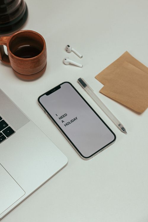 Free A minimalist desk setup with a phone displaying 'I Need a Holiday,' next to a laptop and coffee. Stock Photo