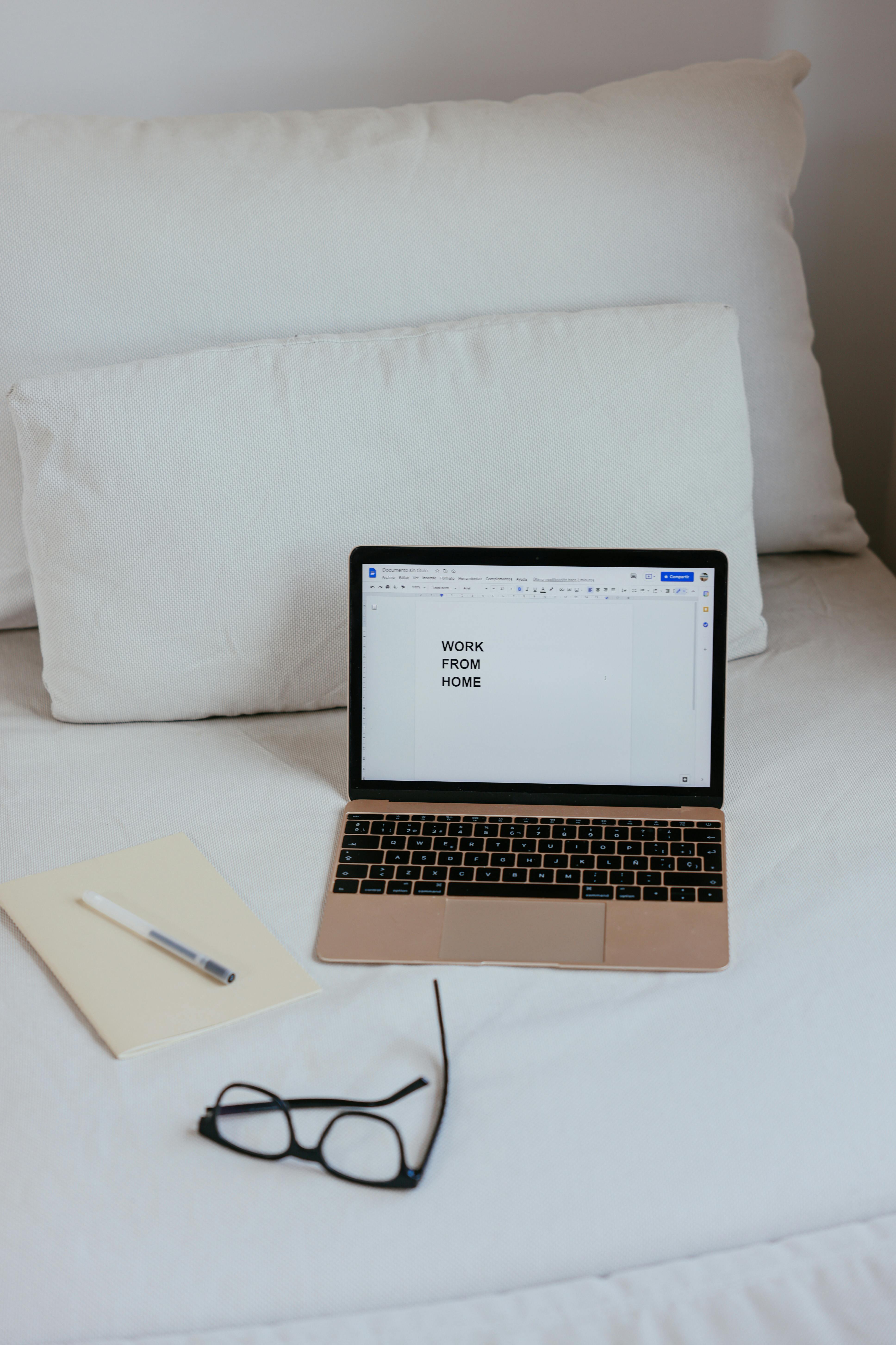 A minimalist work from home setup with a laptop on a white bed, eyeglasses, and notebook.