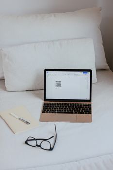 A minimalist work from home setup with a laptop on a white bed, eyeglasses, and notebook.