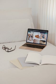 A comfortable bedroom setting with an open laptop, notebooks, and eyeglasses on a bed.