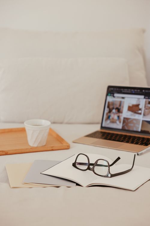 Free A comfortable workspace setup on a bed, featuring a laptop, notebook, and eyeglasses with a cup on a wooden tray. Stock Photo