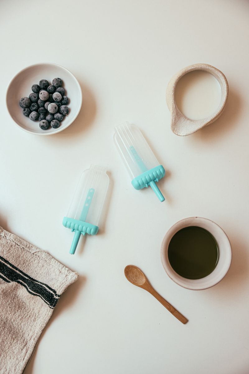 Top-down view of popsicle molds being filled with milk, blueberries and green tea