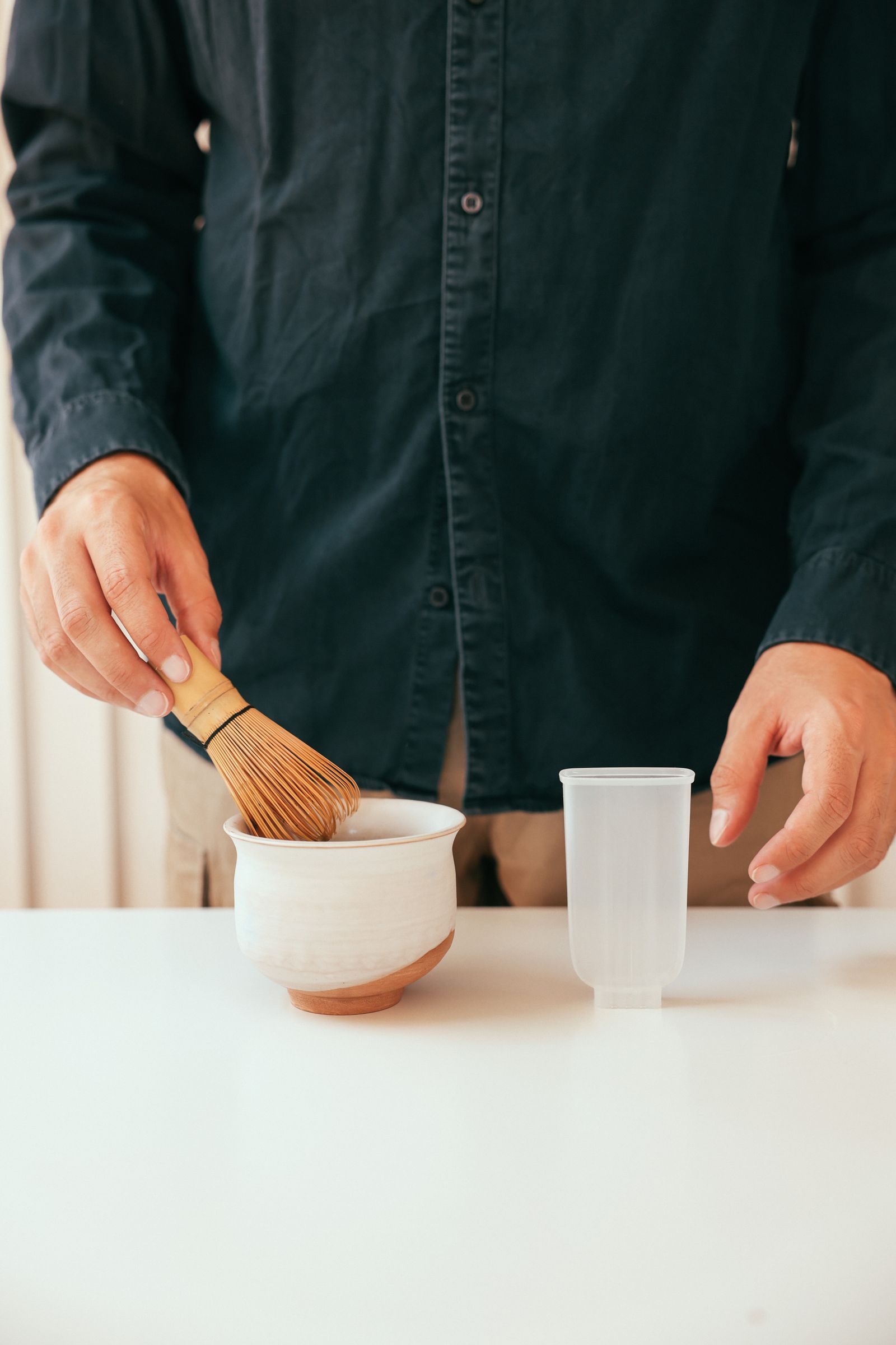 Person moving a bamboo whisk inside a narrow ceramic bowl beside a white cup