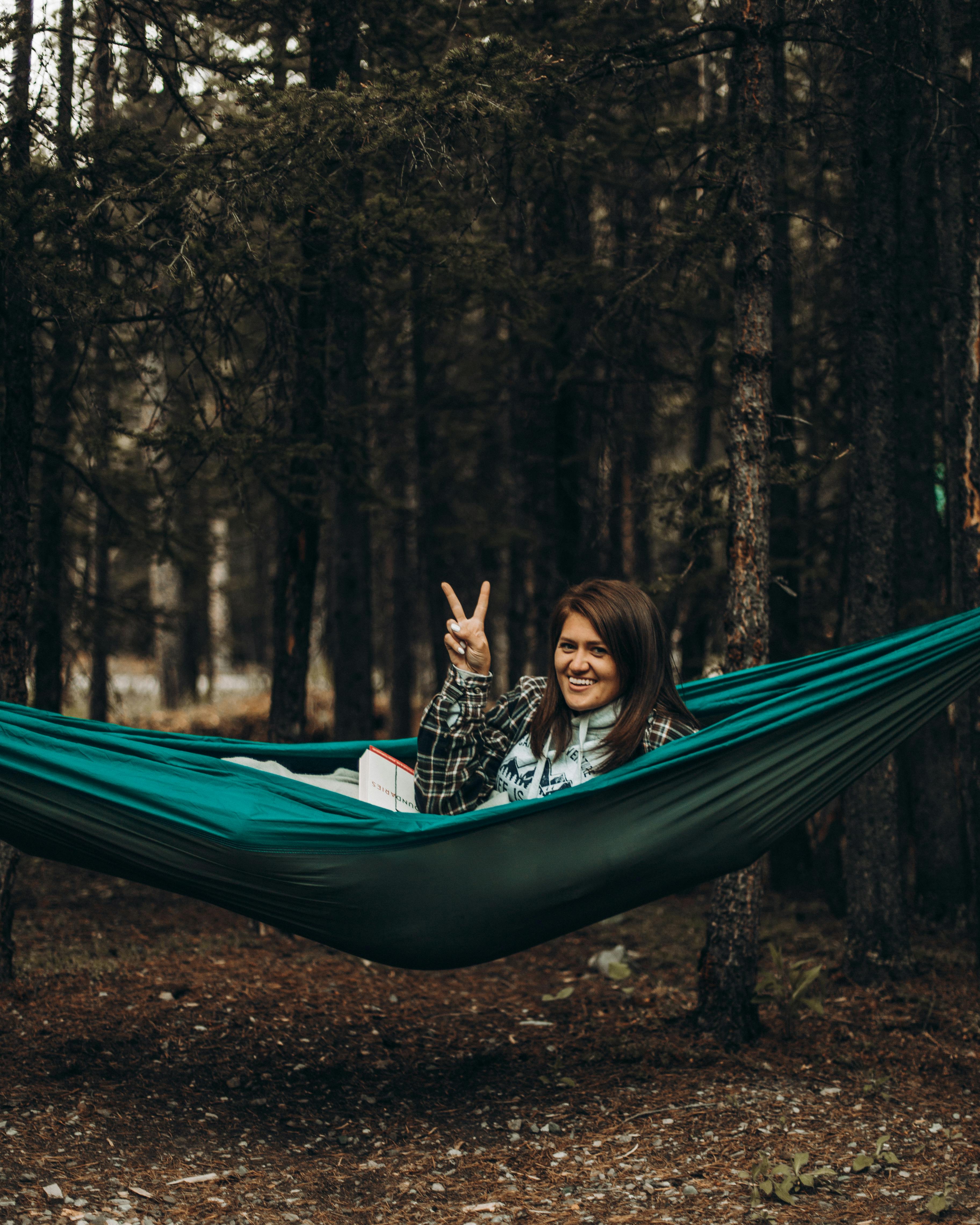 Woman smiles and makes peace sign while relaxing in a hammock in a woodland setting.