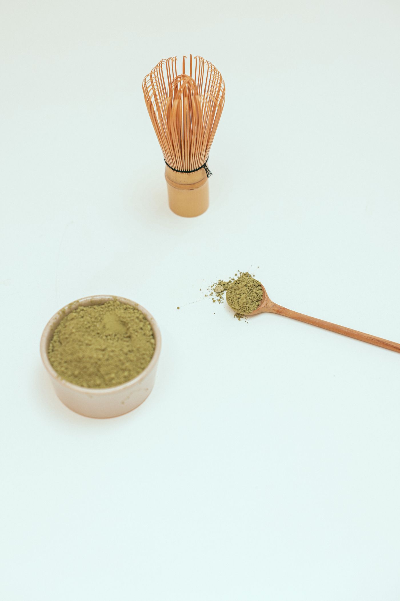 Bamboo whisk beside a bowl of matcha powder and a filled wooden spoon on a pale surface