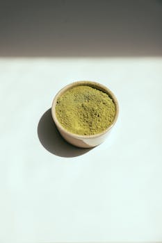 A minimalist ceramic bowl filled with vibrant green matcha powder on a white background.