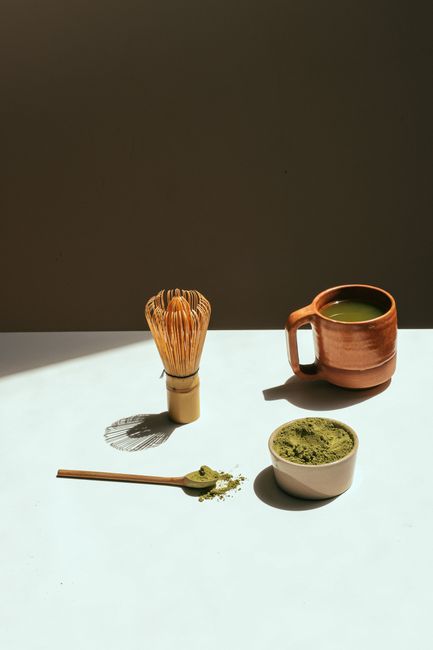 A minimalist still life featuring matcha tea, a bamboo whisk, and tea accessories in warm lighting.