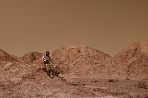 A Man in Space Suit Sitting on a Rock Formation in the Desert