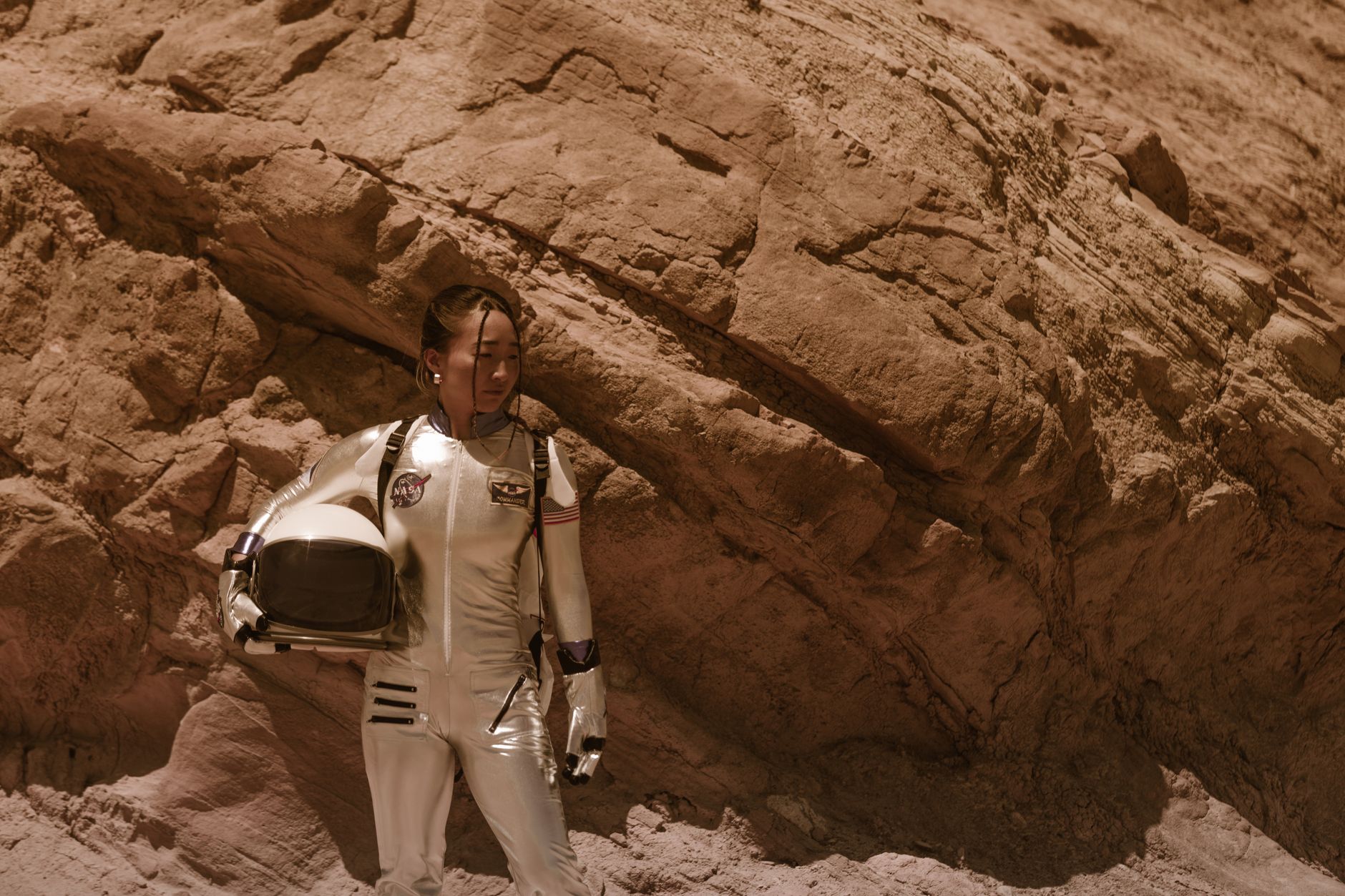 Woman astronaut in space suit standing against desert rock formation reflecting Mars scenery.