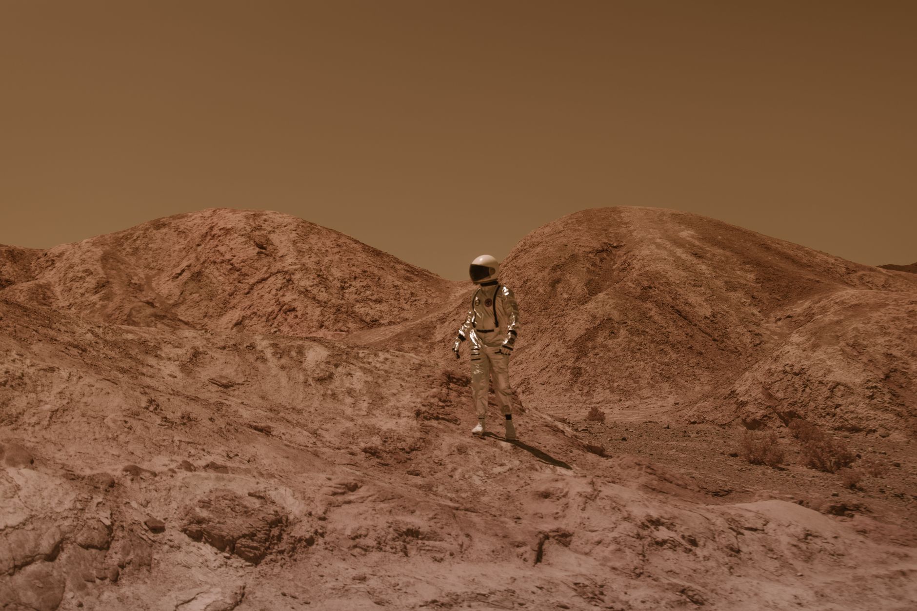 Astronaut in spacesuit explores barren, Mars-like desert terrain under brown sky.