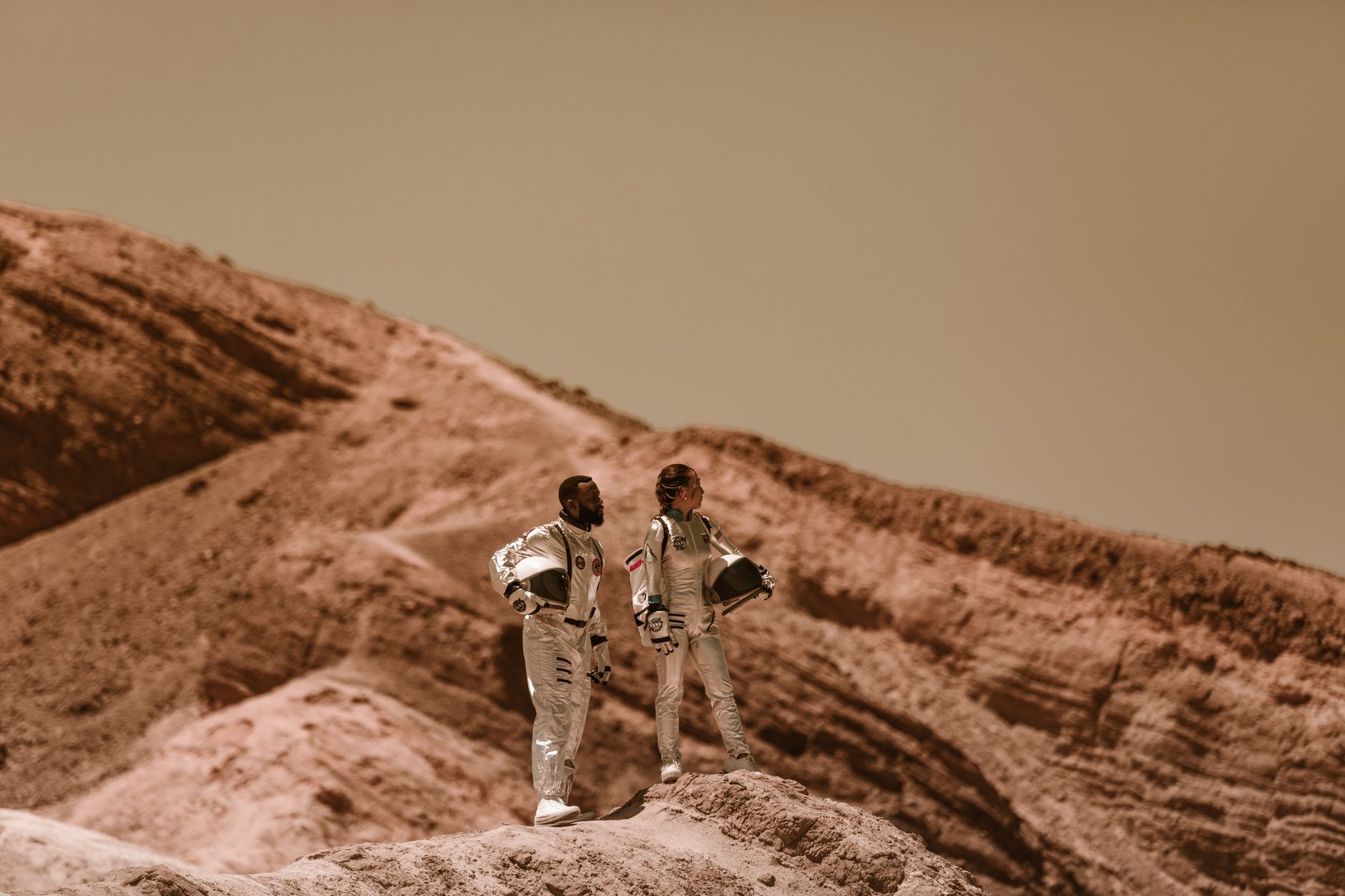Astronauts exploring a desert-like terrain with rugged hills, wearing space suits.