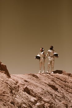 Two astronauts in spacesuits on rocky terrain, resembling Mars exploration.
