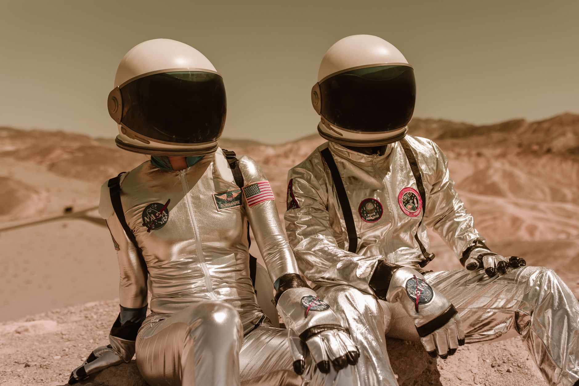 Two astronauts in silver suits and helmets sitting on a Mars-like desert landscape.