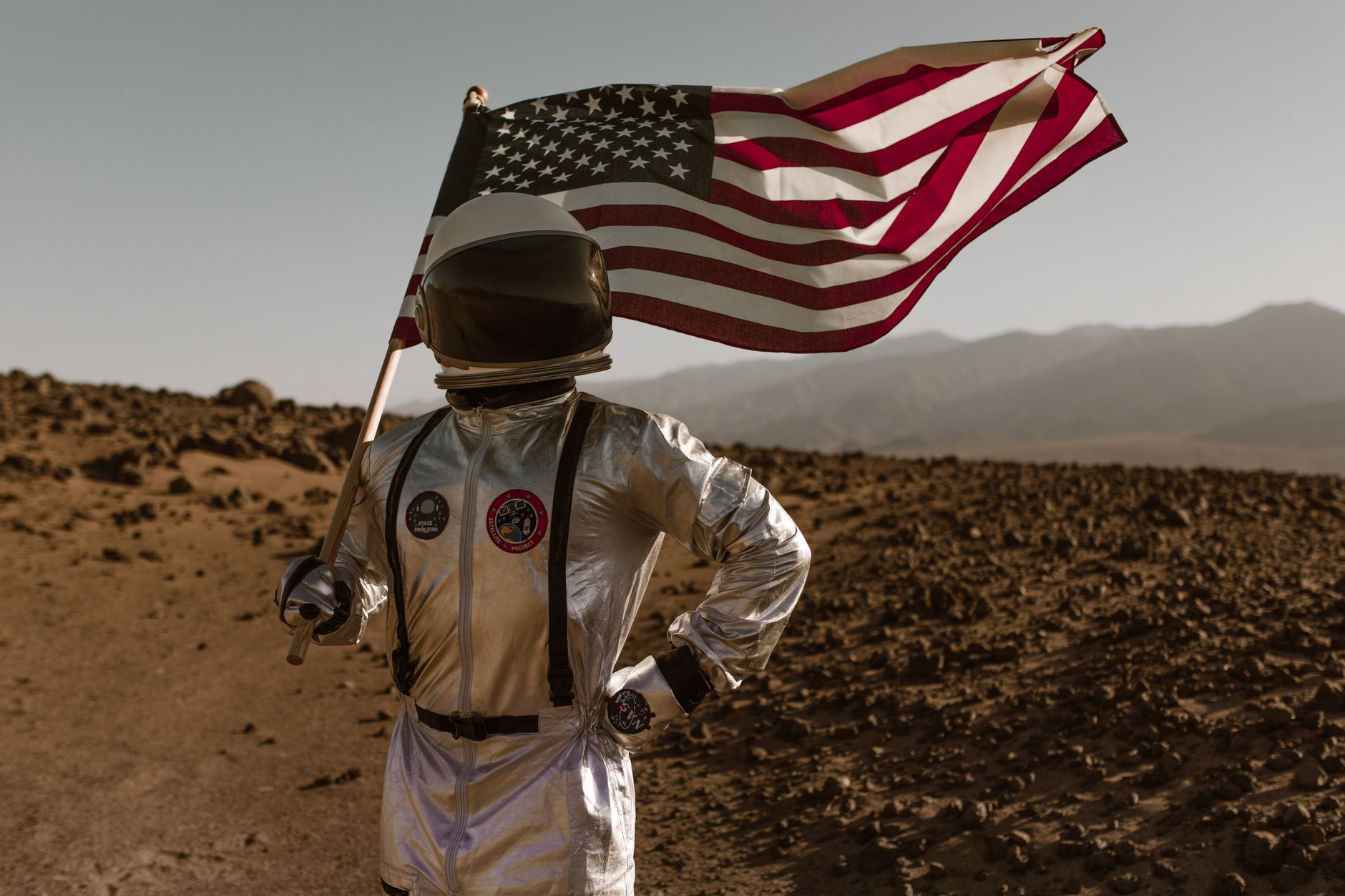 Astronaut in a silver suit holding an American flag, standing on rocky terrain resembling Mars.