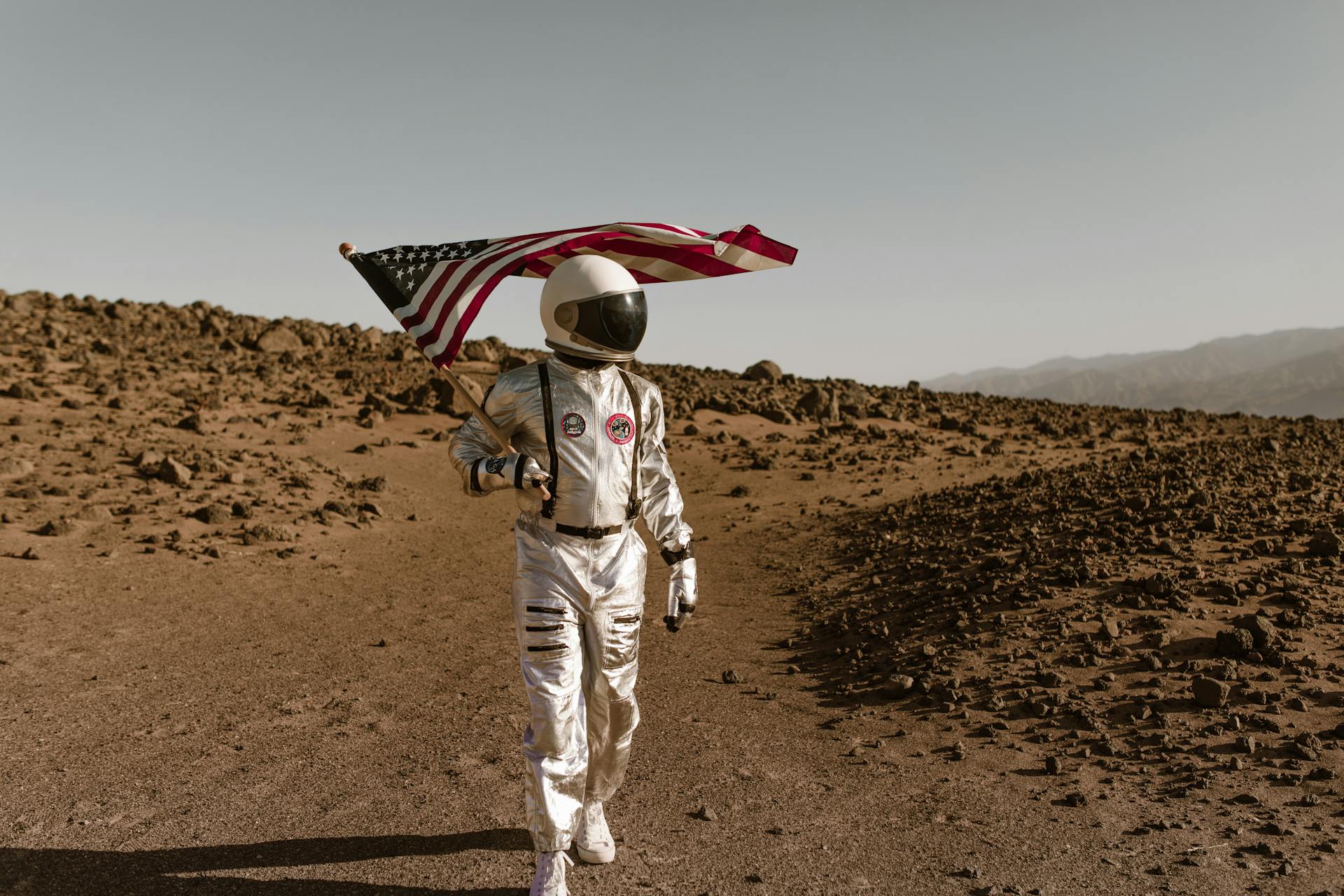 An Astronaut Walking in a Desolate Area while Holding an American Flag